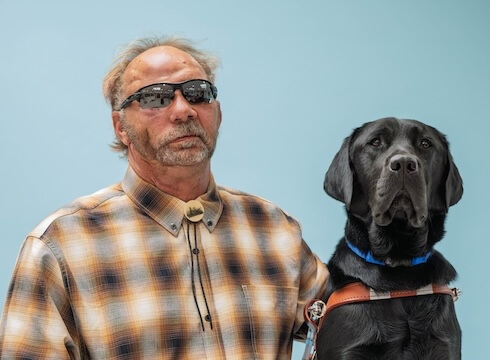 Spencer and black Lab guide dog Commander sit side by side for their team portrait against a pale blue background.