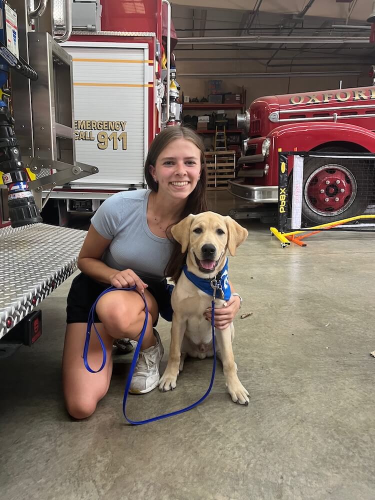 Pup Vernon wears a blue bandana and poses with raiser Megan in a firehouse