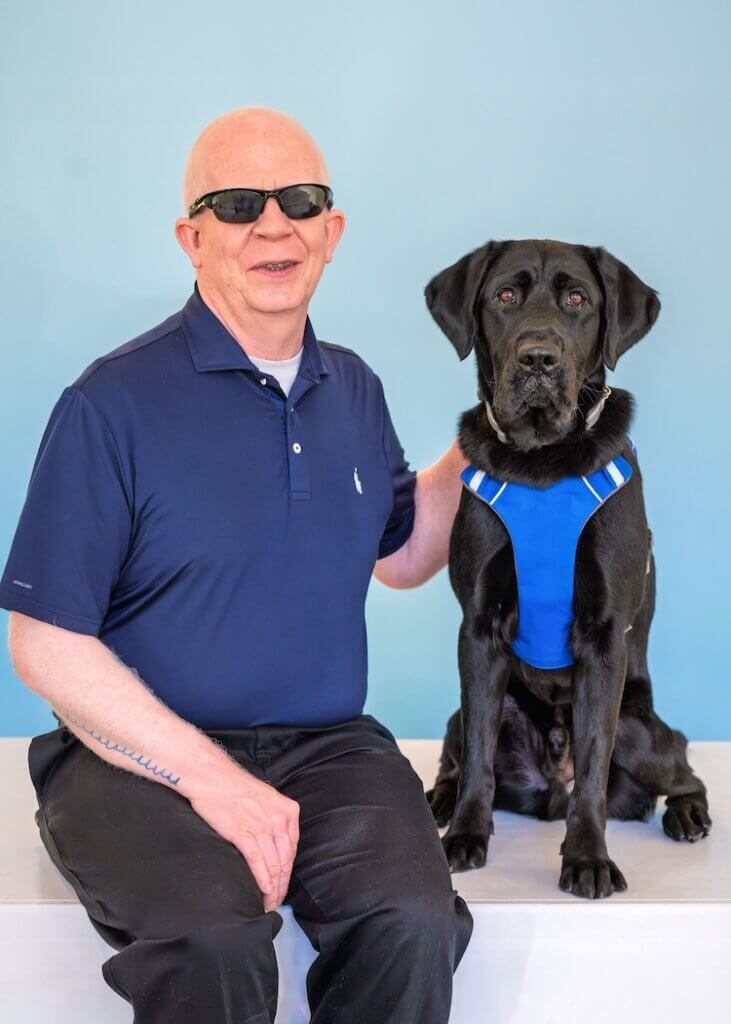 Willie Lab and black Lab guide dog Bono sit side by side for their team portrait against a blue background