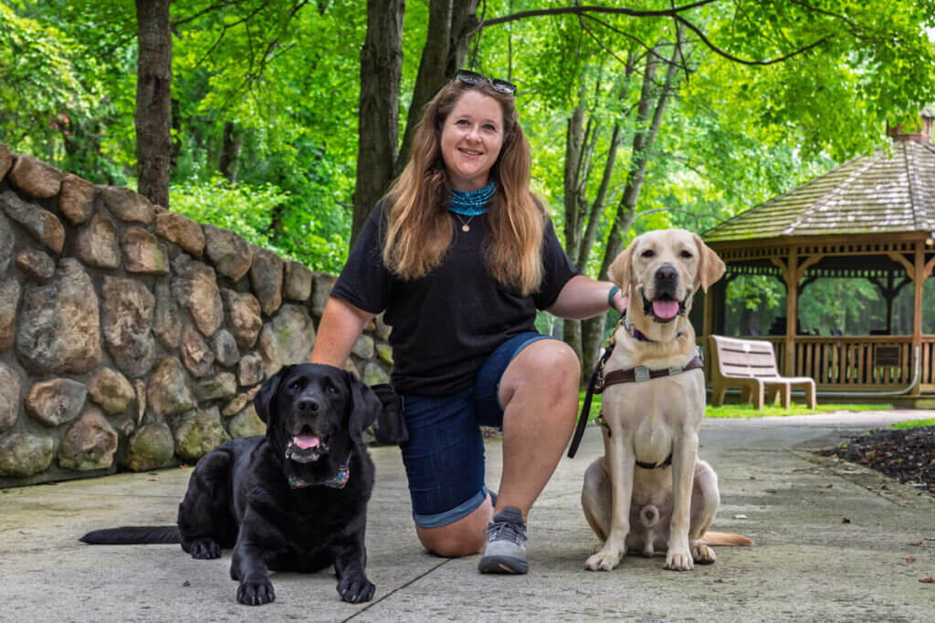 Allie Greenberg, a Guide Dog Mobility Instructor, kneels on the Nature Path at the Guiding Eyes Training School campus with Lopez, her male black Labrador pet dog, lying beside her and Vassar, a yellow Labrador guide dog in training, sitting at her other side.