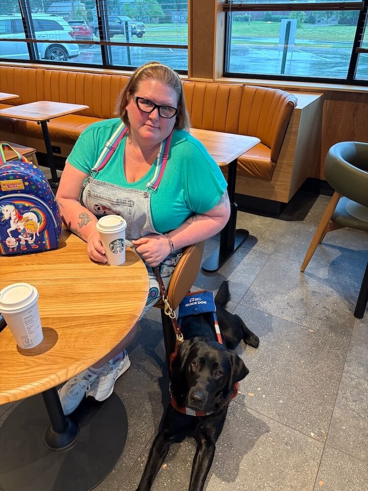 Amanda sits at a coffee shop with her hand on the cup and guide Major resting on the floor