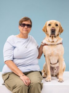 Beth and yellow Lab guide dog Eclipse sit side by side for their team portrait against a light blue background