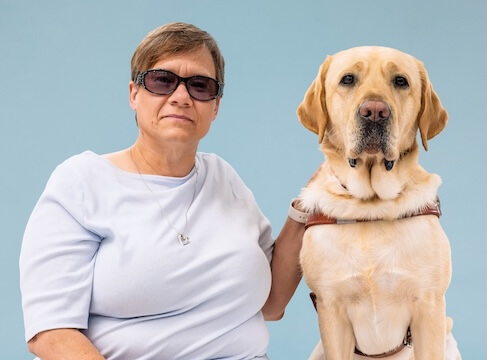 Beth and yellow Lab guide dog Eclipse sit side by side for their team portrait against a light blue background