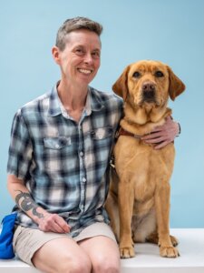 H and yellow Lab guide dog Barben sit side by side for their team portrait against a light blue background