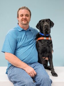 Joe and black Lab guide dog Taco sit side by side for their team portrait against a pale blue background