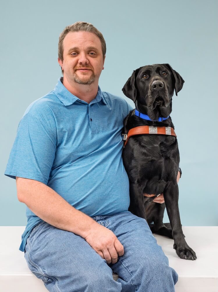 Joe and black Lab guide dog Taco sit side by side for their team portrait against a pale blue background