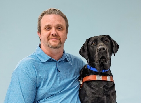 Joe and black Lab guide dog Taco sit side by side for their team portrait against a pale blue background