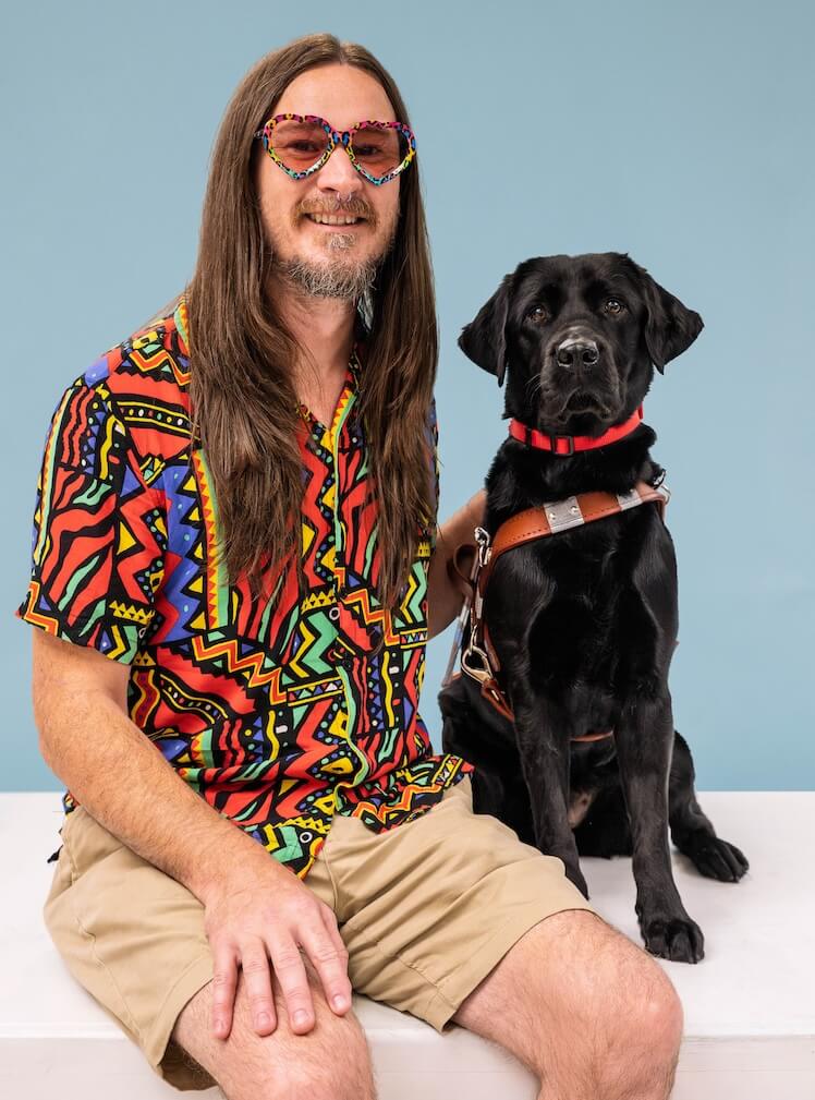 Kevin and black Lab guide dog Yahtzee sit side by side for their team portrait against a pale blue background
