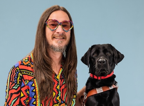 Kevin and black Lab guide dog Yahtzee sit side by side for their team portrait against a pale blue background