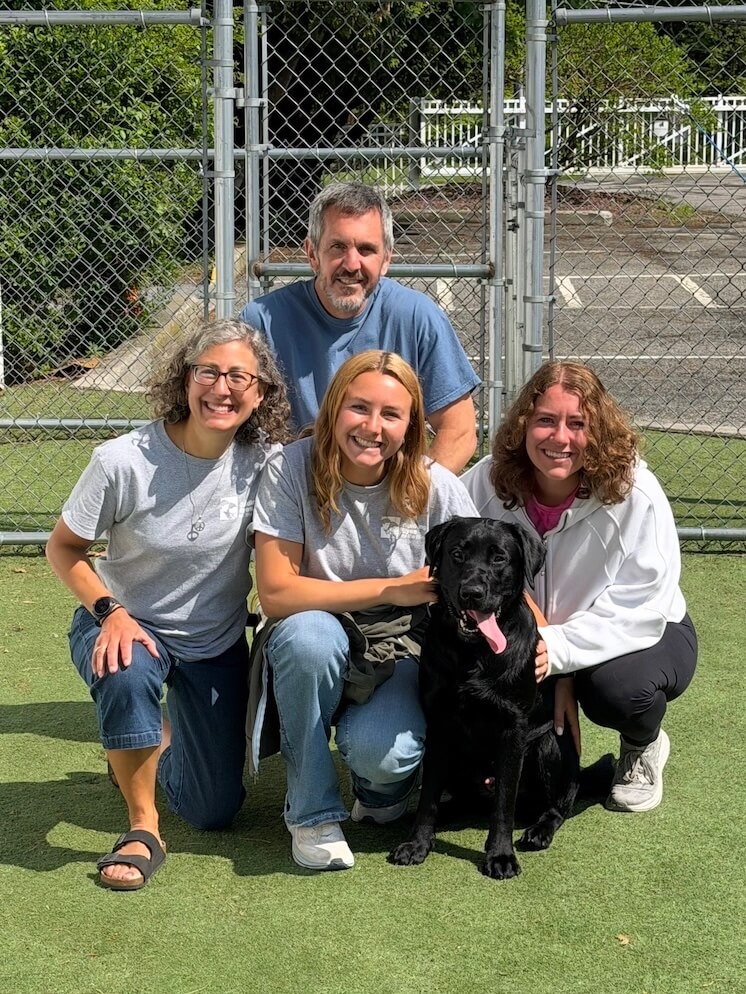 Black Lab Major sits with tongue out as his four raisers gather around him on bent knees