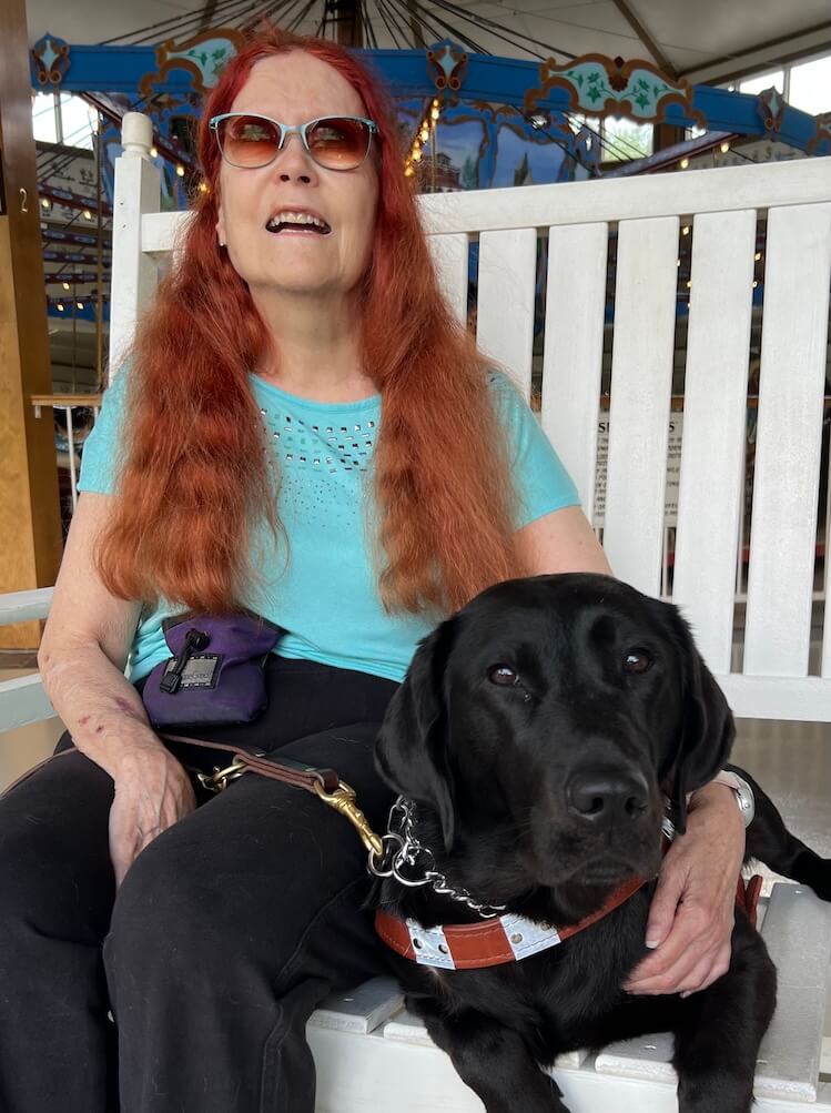 Mary Ann and black Lab guide dog JoJo sit at a bench at the edge of a carousel