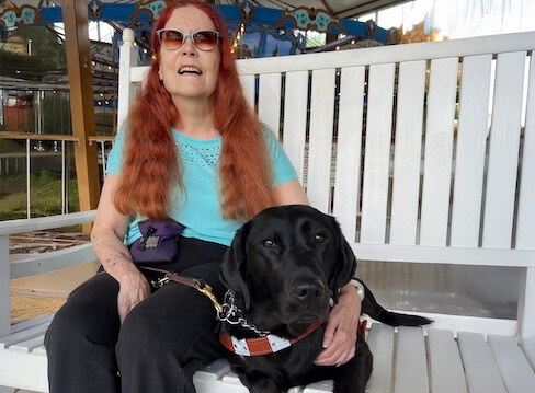 Mary Ann and black Lab guide dog JoJo sit at a bench at the edge of a carousel