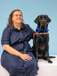Rachel and black Lab guide dog Vic sit side by side for their team portrait against a pale blue background