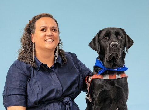 Rachel and black Lab guide dog Vic sit side by side for their team portrait against a pale blue background