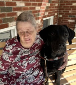 Rita and black Lab guide dog Julie sit on a wooden bench in the shade of a brick building