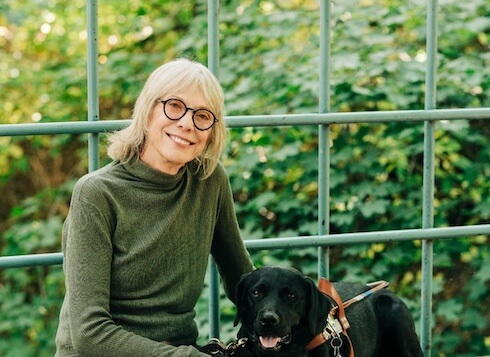 Deni sits on a low stone bench with guide dog Hildy against a backdrop of lush greenery beyond a large window -Kristine_Paulsen_Photography