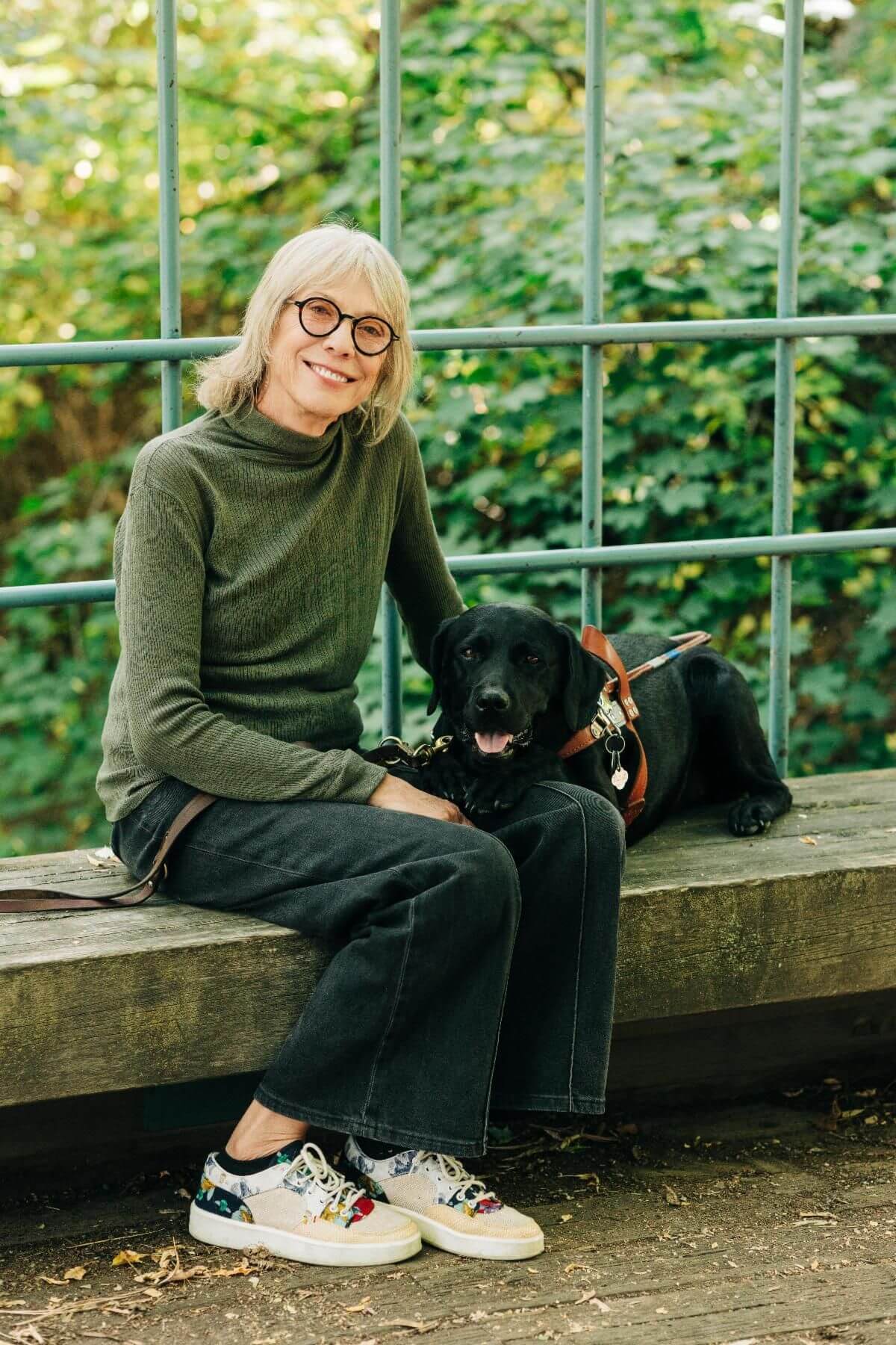 Deni sits on a low stone bench with guide dog Hildy against a backdrop of lush greenery beyond a large window -Kristine_Paulsen_Photography