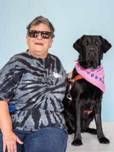 Elizabeth and black Lab guide dog Octavia sit against a pale blue background for their team portrait