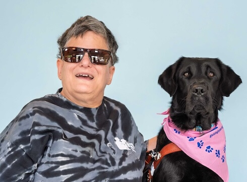 Elizabeth and black Lab guide dog Octavia sit against a pale blue background for their team portrait