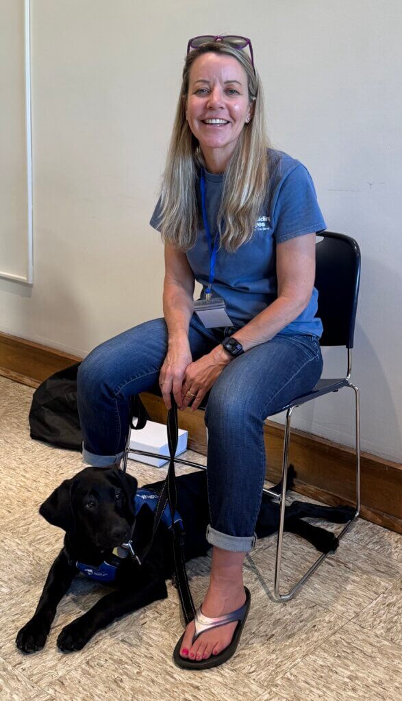Jennifer Thurman, Regional Puppy Instructor, sits in a chair at a conference with Etta, a black Lab puppy in the Guiding Eyes Puppy Raising program, resting on the floor beside her feet. Etta wears her Guiding Eyes puppy vest while Jennifer holds her leash.