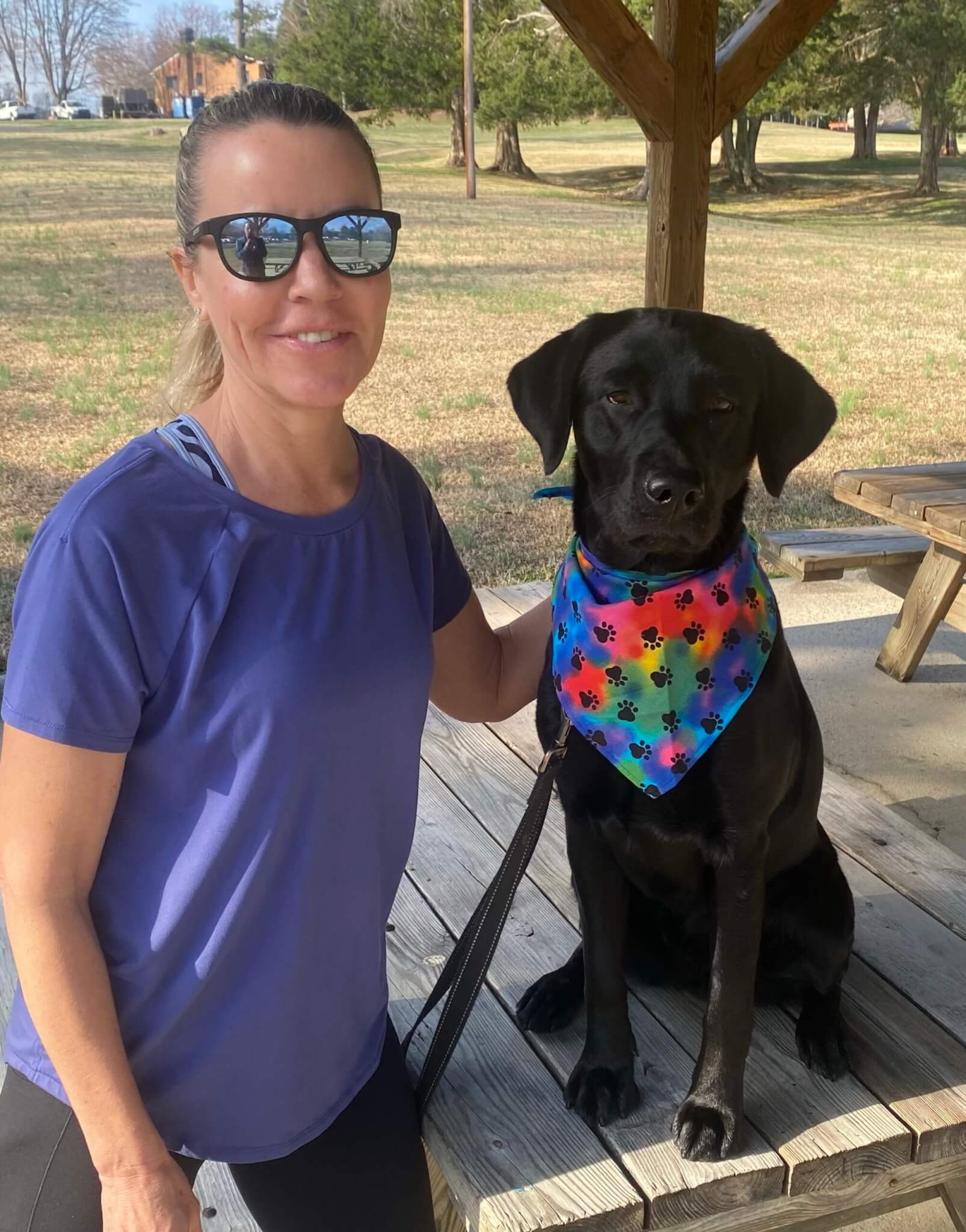 Jennifer Thurman, Regional Puppy Instructor, sits beside Etta, a black Lab puppy in the Guiding Eyes Puppy Raising program, at an outdoor picnic table. Etta wears a colorful tie-dye bandana patterned with black paw prints.