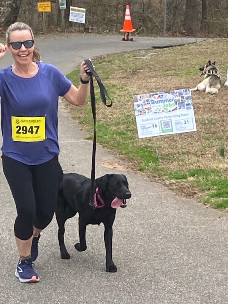 Jennifer Thurman, Regional Puppy Instructor, jogs beside Etta, a black Lab puppy in the Guiding Eyes Puppy Raising program, during a 5K. Jennifer smiles and raises one arm while holding Etta’s leash, and Etta runs alongside her with her tongue out.