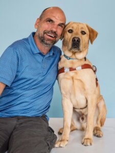 Jim and yellow Lab guide dog Keanu sit side by side for their team portrait against a light blue background