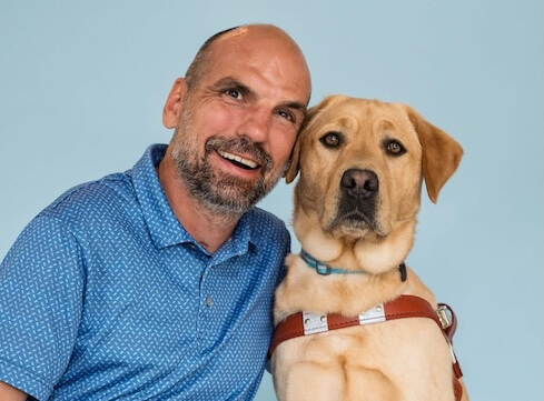 Jim and yellow Lab guide dog Keanu sit side by side for their team portrait against a light blue background