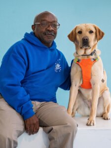 Joe and yellow Lab guide dog Garden sit side by side for their team portrait against a light blue background