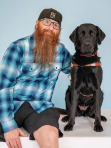 Joe and black Lab guide dog Shanna sit side by side for their team portrait against a light blue background