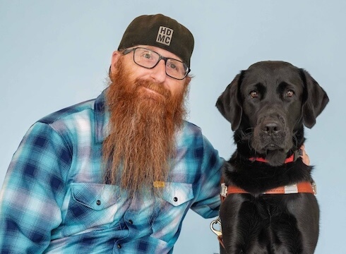 Joe and black Lab guide dog Shanna sit side by side for their team portrait against a light blue background