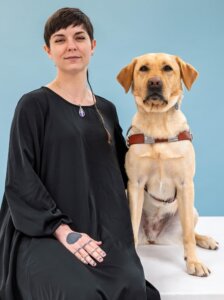 Autumn and yellow Lab guide dog Angie sit side by side for their team portrait against a light blue background
