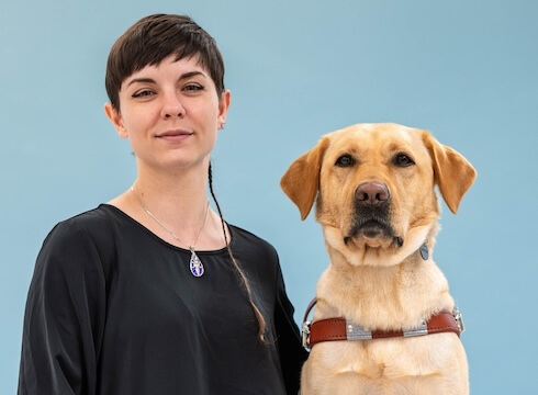 Autumn and yellow Lab guide dog Angie sit side by side for their team portrait against a light blue background