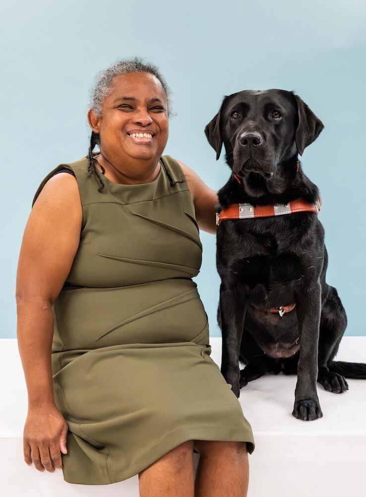 Danielle and black Lab guide dog Natasha sit side by side for their team portrait against a light blue background
