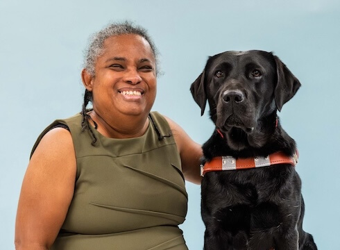 Danielle and black Lab guide dog Natasha sit side by side for their team portrait against a light blue background