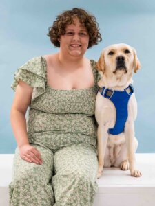 Franny and yellow Lab guide dog Tatum sit side by side for their team portrait against a light blue background