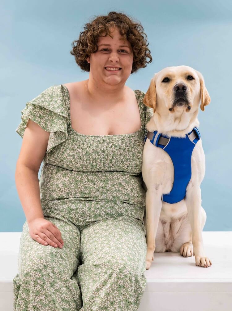 Franny and yellow Lab guide dog Tatum sit side by side for their team portrait against a light blue background
