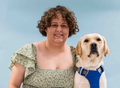 Franny and yellow Lab guide dog Tatum sit side by side for their team portrait against a light blue background