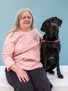 Gail and black Lab guide dog Mimi, doing an endearing head tilt, sit side by side for their team portrait against a light blue background