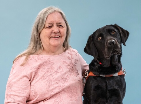 Gail and black Lab guide dog Mimi, doing an endearing head tilt, sit side by side for their team portrait against a light blue background