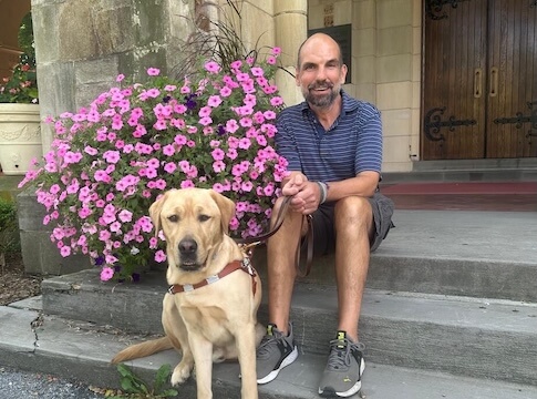 Handler Jim and guide dog Keanu sit on the steps of a grand entrance next to a huge pot of pink flowers