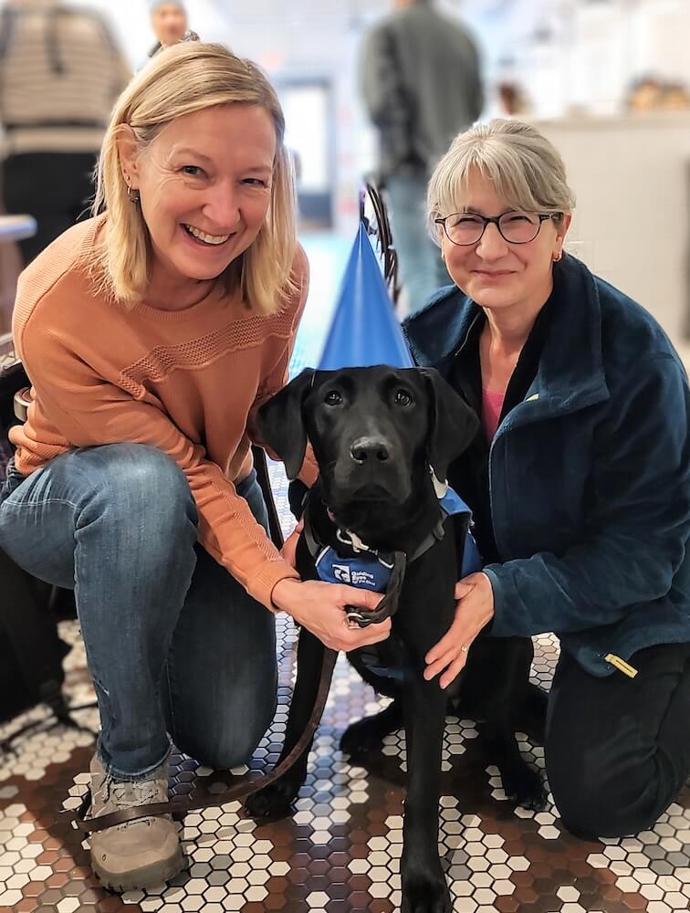 Black Lab pup Natasha with raisers Cheryl and Naomi kneeling on either side