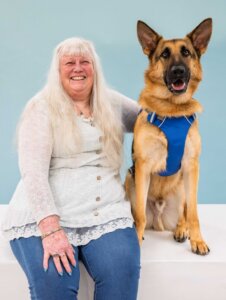 Pam and German Shepherd guide dog Usher sit for their team portrait against a light blue background