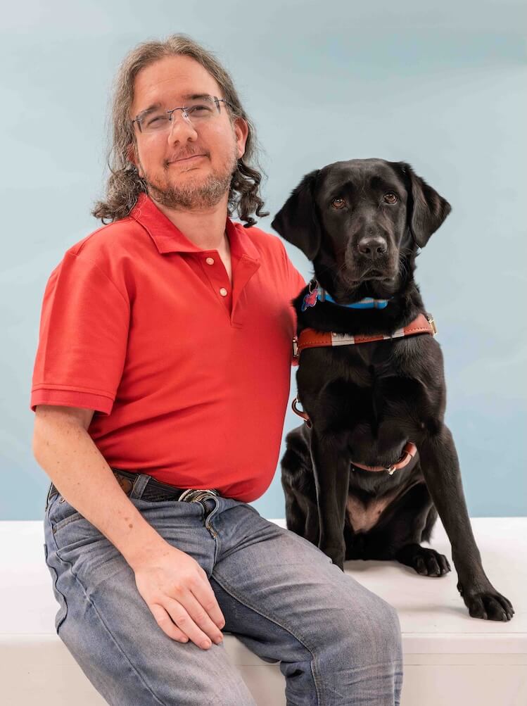 Tom and black Lab guide dog Nellie sit side by side for their team portrait against a light blue background
