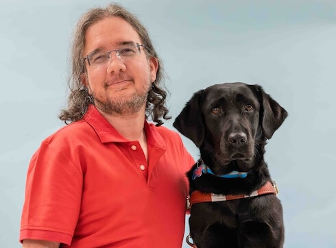 Tom and black Lab guide dog Nellie sit side by side for their team portrait against a light blue background