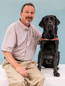 Wade and black Lab guide dog Keaton sit side by side for their team portrait against a light blue background