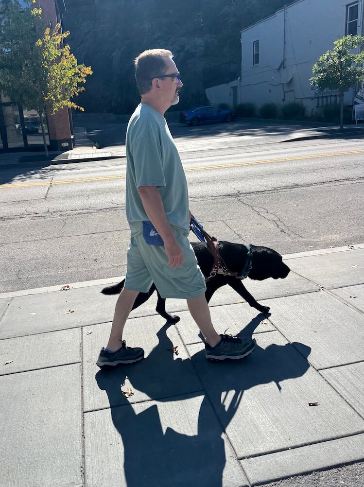 Wade and guide Keaton walk safely down a sidewalk with buildings seen on the other side of the street